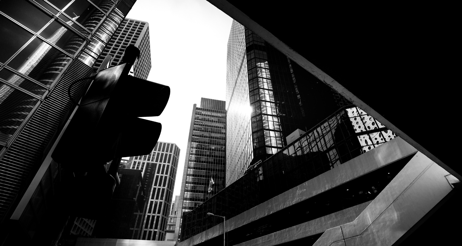 Image shows a city from the street, looking up at the buildings under an overpass. The sides of buildings and the tops of skyscrapers are seen. There is a traffic light in view. The image is in black and white. 