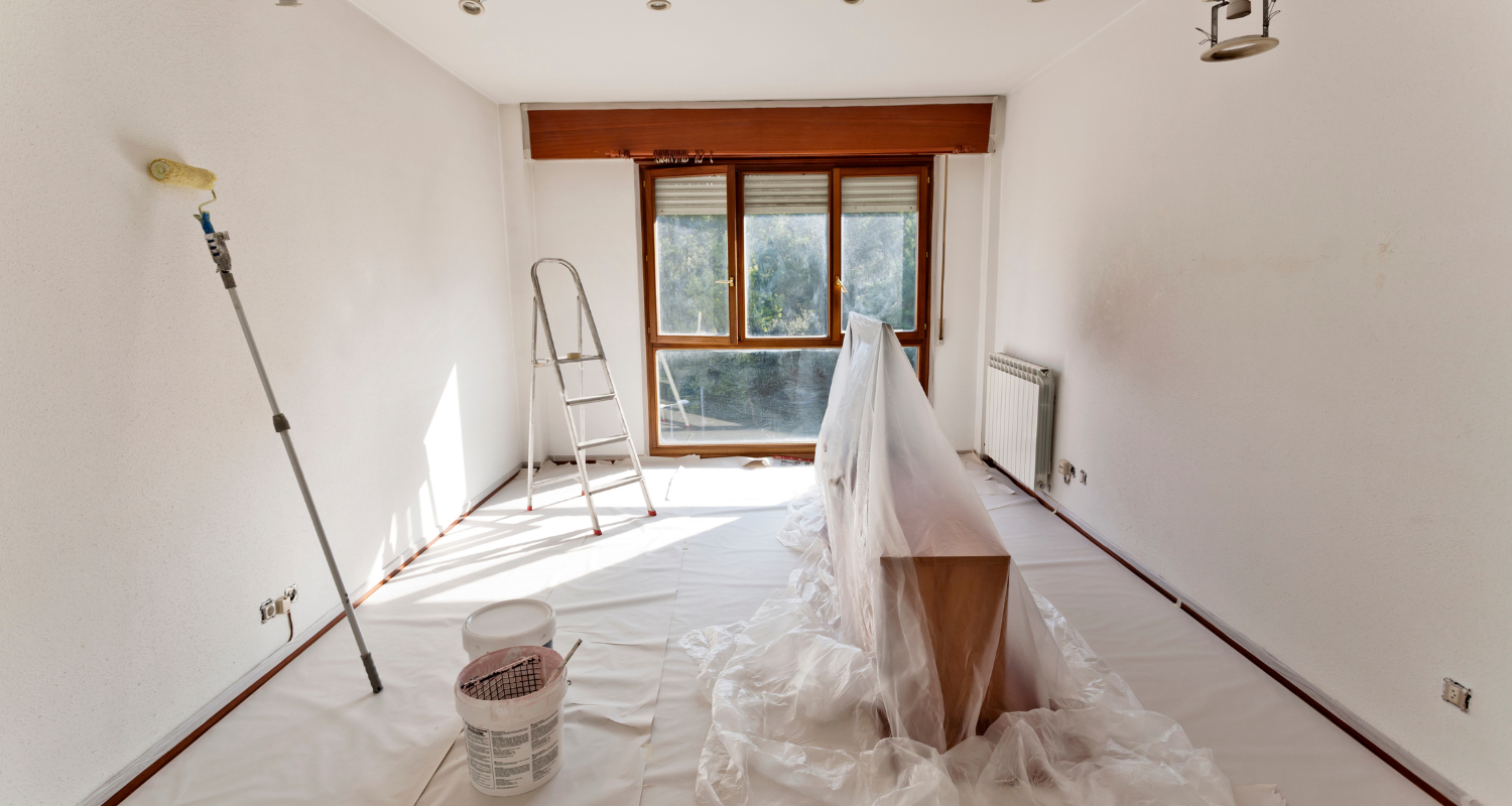 A room with white walls and a window at the end. Sunlight is shining through the window. Painting equipment is in the room, and plastic is set down on the floor and over a desk. This may be an example of what to do when you've accidentally used exterior paint indoors. 