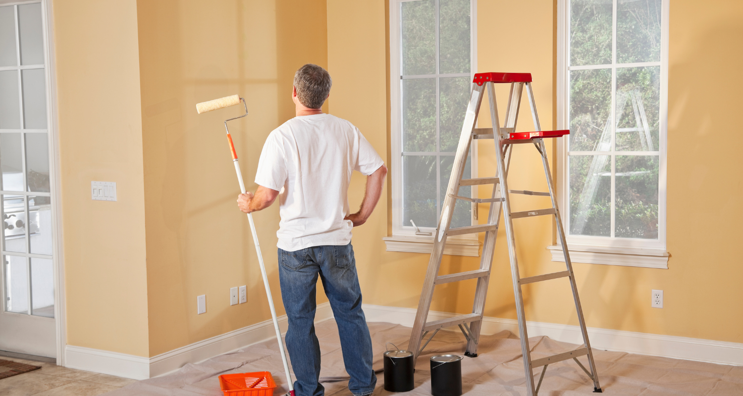 A man stands in front of a peach colored wall. He holds a paint roller in his hand. A ladder is open next to him.