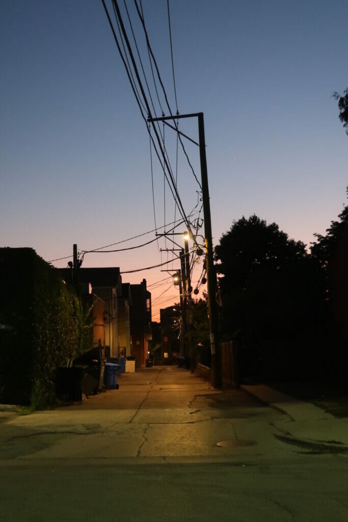 The sun sets above a Chicago alleyway. Light poles and garbage cans are seen. 